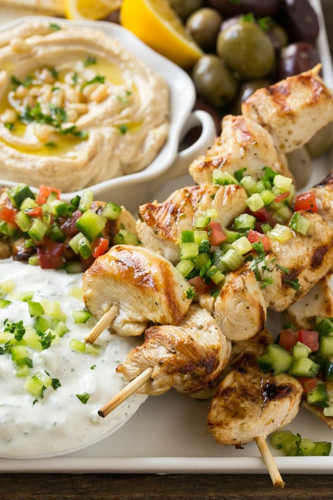 Greek Chicken Souvlaki Recipe | Easy Greek Chicken | Chicken Souvlaki | Greek Chicken Skewers | Easy Greek Chicken Skewers #chicken #kabobs #glutenfree #keto #lowcarb #grilling #sraye A tray of chicken souvlaki kabobs with olives, hummus, pita bread and yogurt sauce.