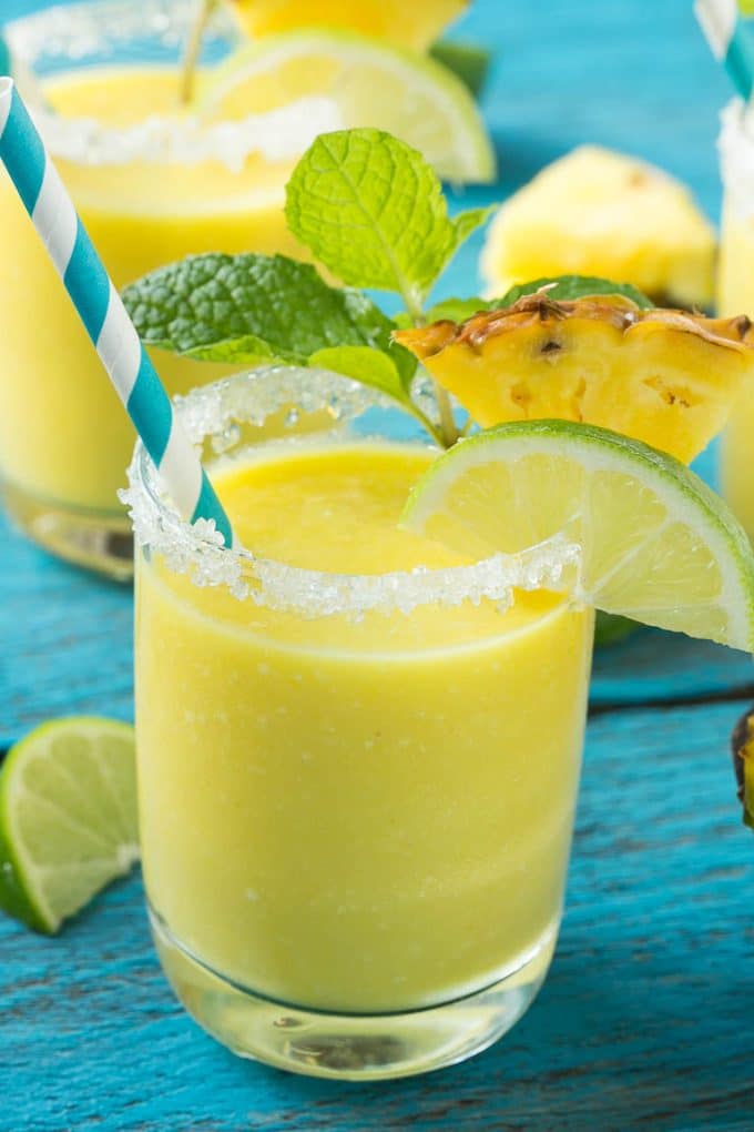 Pineapple Coconut Smoothie Recipe |Tropical Smoothie | Pineapple Smoothie #smoothie #pineapple #coconut #healthy #cleaneating #sraye A cup of pineapple smoothie with garnishes of lime, pineapple wedges and mint sprigs.