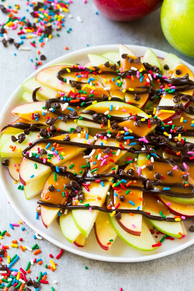 Apple Nachos Recipe | After School Snack | Caramel Apples #apple #apples #caramelapple #snack #dessert #sraye A plate of sliced apple nachos topped with caramel and chocolate sauces, rainbow sprinkles and mini chocolate chips