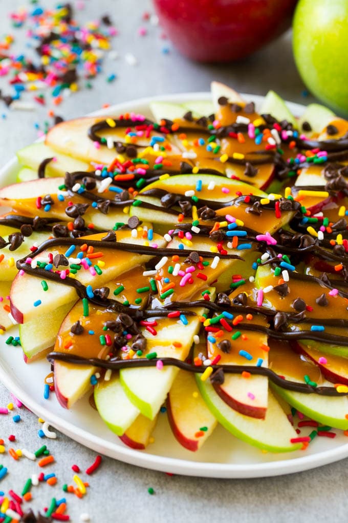 Apple Nachos Recipe | After School Snack | Caramel Apples #apple #apples #caramelapple #snack #dessert #sraye A plate of red and green sliced apples drizzled with caramel and chocolate, then topped with rainbow sprinkles and chocolate chips.