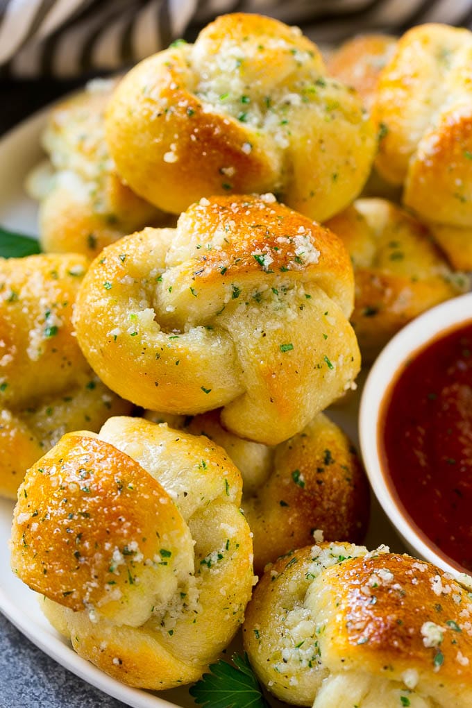 Garlic Knots Recipe | Garlic Rolls | Garlic Butter Rolls | Garlic Bread Recipe #garlic #garlicbread #garlicknots #breadsticks #sraye A plate of garlic knots slathered in butter next to a cup of marinara sauce.