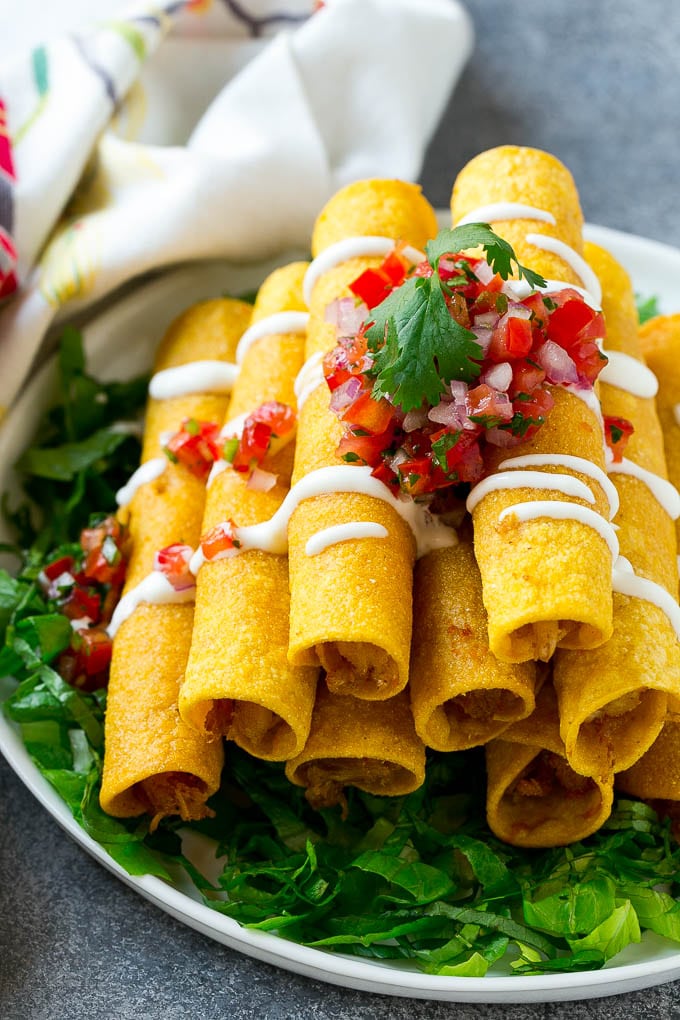 Chicken Taquitos Recipe | Baked Chicken Taquitos | Fried Chicken Taquitos | Mexican Appetizer | Chicken Tortillas #taquitos #chicken #appetizer #tortillas #mexicanfood #sraye A stack of chicken taquitos with a drizzle of sour cream and pico de gallo topping.
