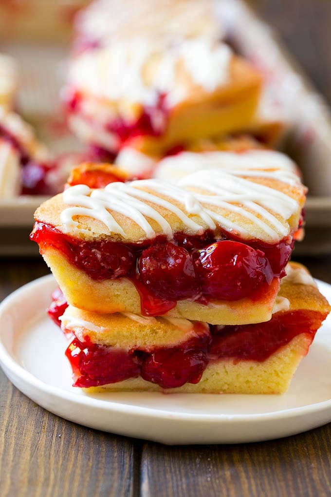 Cherry Bars Recipe | Cherry Pie Bars | Cherry Cookies | Bar Cookies #cookies #bars #cherry #baking #dessert #sweets #sraye Two cherry bars on a plate with a pan of bars in the background.