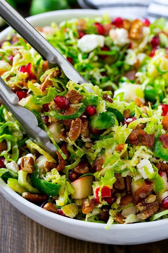 Brussels Sprout Salad Recipe | Shredded Brussels Sprouts Salad | Fall Salad #salad #brusselssprouts #bacon #thanksgiving #pomegranate #apple #dinner #sraye A bowl of brussels sprout salad with tongs picking some up out of the bowl.