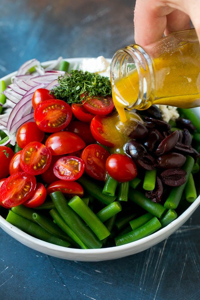Green Bean Salad Recipe | String Bean Salad | Chopped Salad #greenbeans #tomatoes #olives #salad #cleaneating #lowcarb #glutenfree #sraye Dressing being poured over veggies like cherry tomatoes, olives and onions.
