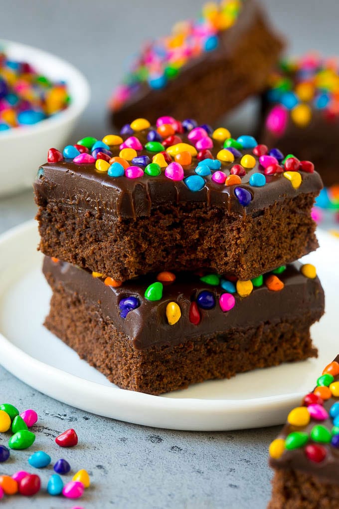 Cosmic Brownies Recipe | Cocoa Brownies | Frosted Brownies #brownies #chocolate #dessert #sraye #sprinkles A stack of cosmic brownies on a plate with a bite taken out of one.