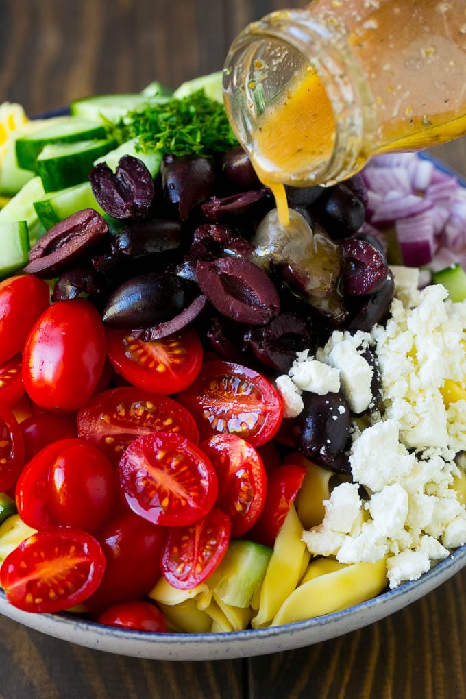 Greek Tortellini Salad Recipe | Pasta Salad | Tortellini Salad | Greek Salad #greek #tortellini #pasta #salad #cucumbers #olives #dinner #sraye Salad dressing being poured over a bowl of tortellini and vegetables.