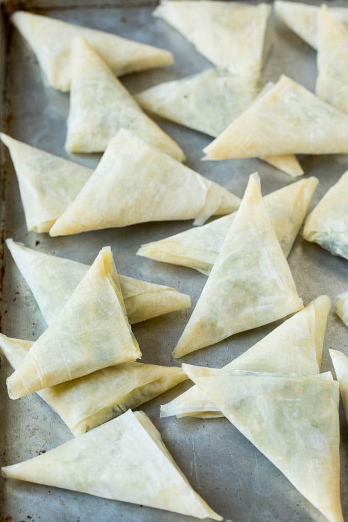 Spanakopita Recipe #appetizer #snack #spinach #cheese #sraye Folded stuffed phyllo triangles on a sheet pan.