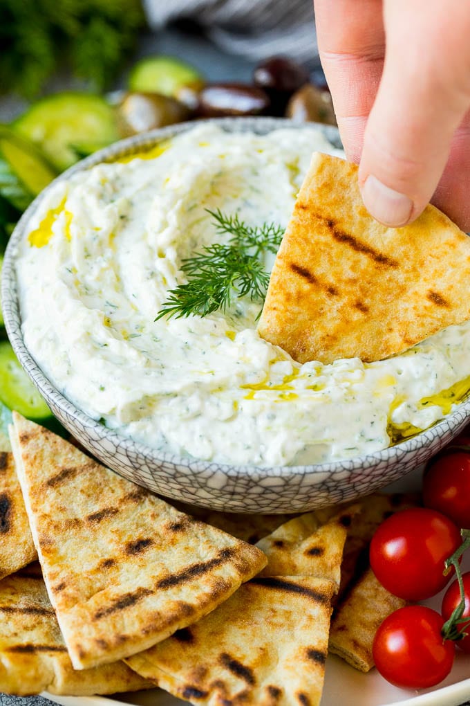 The best homemade tzatziki sauce with cucumber and herbs mixed into Greek yogurt. #appetizer #sraye A hand dipping pita bread into a bowl of tzatziki.