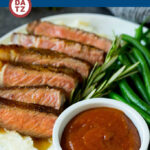 A bowl of steak sauce on a plate with steak, mashed potatoes and green beans.